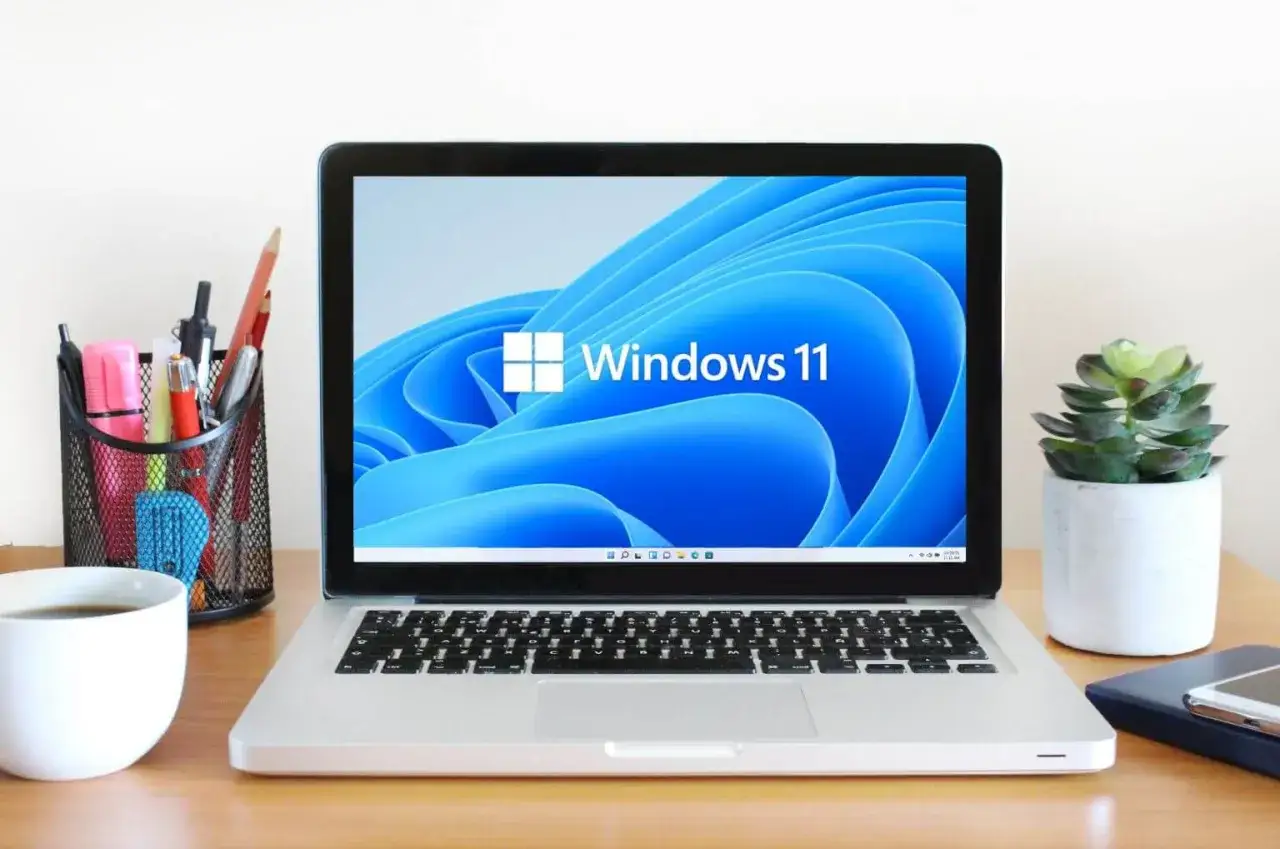 Laptop z ekranem powitalnym Windows 11 na biurku, obok kubek kawy i doniczka z rośliną. Nawet stary komputer może działać z Windows 11.